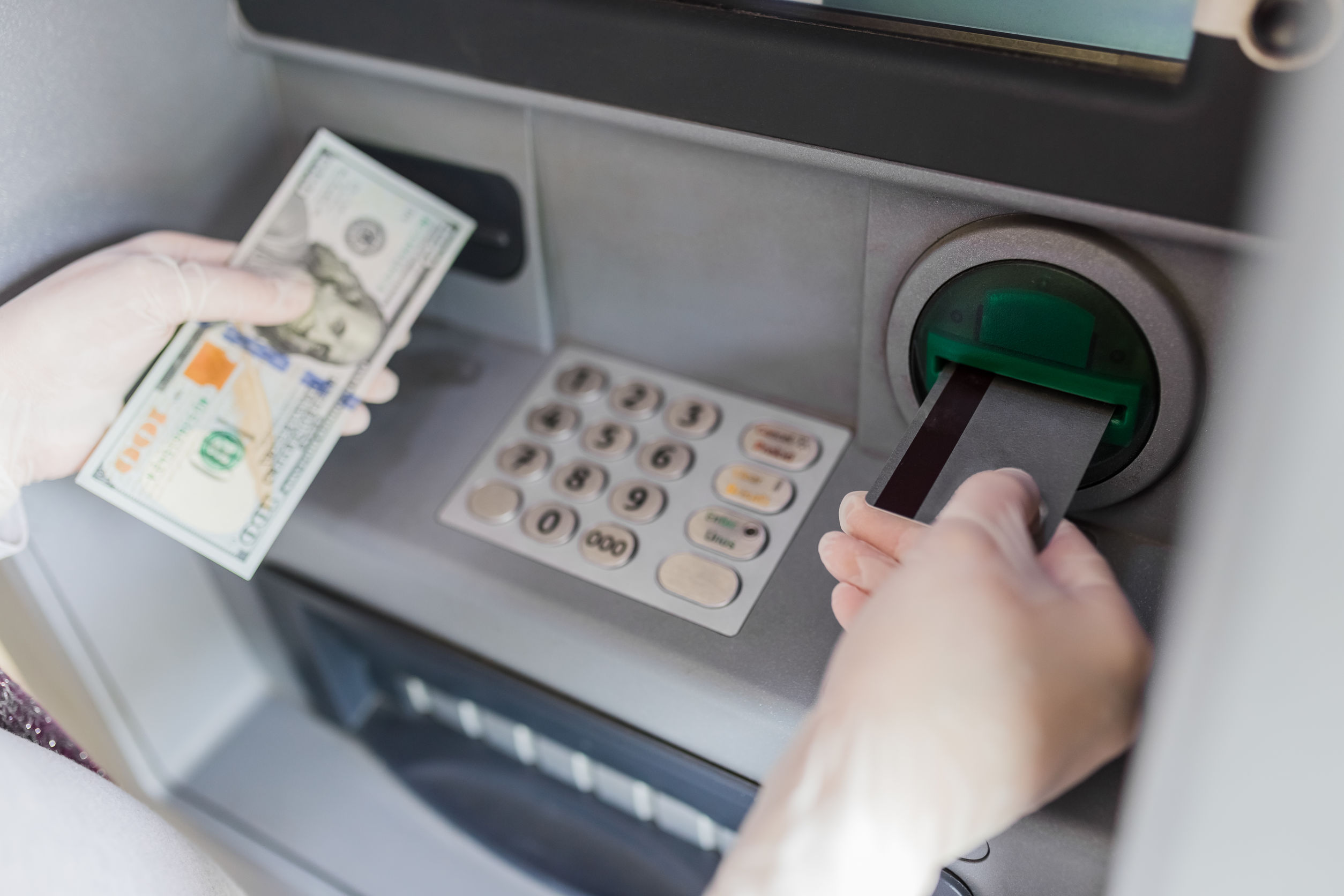 Hands in protective surgical gloves holding one hundred dollar bill and using credit card at atm banking device. Covid 19 concept.